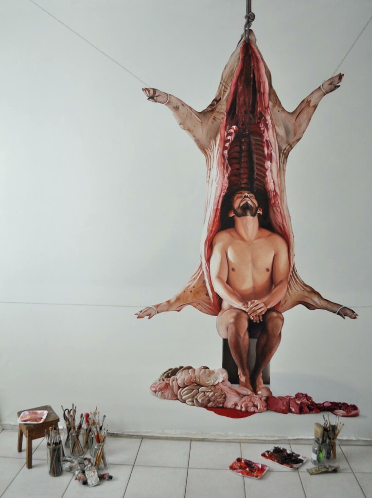 naissance de l'artiste. Fábio Magalhães assis dans une carcasse d'animal pendue dont les viscères sont étalées sur le sol, à côté du matériel de peinture